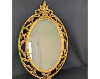 May include: Oval-shaped gold-colored mirror with ornate scrollwork and a decorative top. The mirror's frame has a detailed, vintage design. The mirror is a decorative wall hanging.