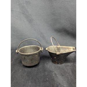May include: Two small, dark metal buckets with arched handles. One is a simple round bucket, and the other has a spout. The buckets are likely made of cast iron and have a rustic, antique appearance. They sit on a dark surface.