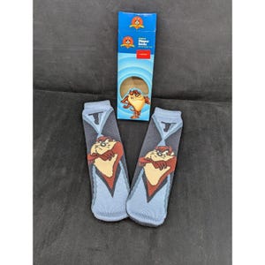 May include: A pair of blue slipper socks featuring the Tasmanian Devil cartoon character. The socks have a gray diamond pattern and a brown and tan Taz graphic. The packaging box is blue and features the Looney Tunes logo.