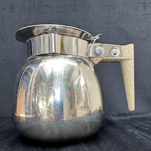 May include: A stainless steel coffee pot with a rounded body and a lid. The handle is a light tan color and is attached to the pot with two screws. The pot has a shiny, reflective surface.