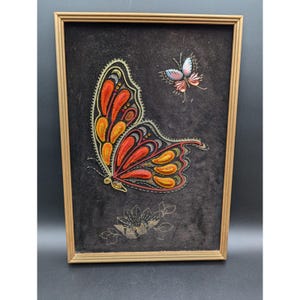 May include: Framed artwork featuring a large butterfly with orange, red, and yellow wings, and a smaller butterfly in pink and blue. The butterflies are embroidered on a dark gray background, with a gold-colored frame.