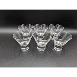 May include: Set of six clear glass cocktail glasses. Each glass has a flared bowl and a detailed etched floral design. The glasses are arranged on a dark surface, showcasing their elegant shape and clarity.