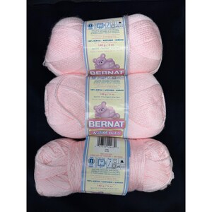 May include: Three skeins of light pink Bernat Softee Baby yarn stacked vertically. Each skein features a label with a teddy bear illustration, the text Bernat Softee Baby, 100% acrylic, and a weight of 140 g / 5 oz. The yarn is a light weight 3.