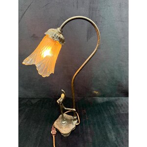 May include: A vintage-style table lamp with a bronze finish. The lamp features a curved arm supporting a flower-shaped, amber-colored glass shade. A small figurine of a dancer is at the base. The lamp is plugged into an electrical outlet.