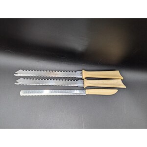 May include: Three bread knives with serrated blades and tan handles are displayed against a dark background. The blades are made of stainless steel. The handles are a solid, neutral color. The knives are designed for slicing bread.