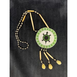 May include: A necklace featuring a circular pendant with white, green, and black seed beads arranged in a turtle pattern. The necklace includes long, thin, cream-colored cylindrical beads and dark metallic spherical beads on a corded strand.