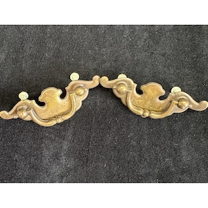May include: Two antique brass drawer pulls with a decorative, curved design. Each pull features a central, ornate shape with rounded details and small screw posts. The brass has a warm, aged patina, suggesting vintage hardware.