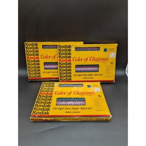 May include: Three yellow boxes of Kodak Color of Christmas lights. Each box features the Kodak logo and text that reads "100 Light Extra Bright Add-A-Set Indoor-Outdoor". The boxes have a red, white, and green zig-zag pattern.