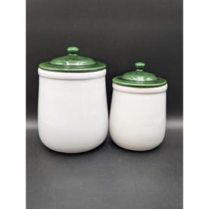 May include: Two white ceramic canisters with smooth, rounded bodies and matching dark green lids. Each lid features a small, centered knob handle. The canisters are cylindrical with a slight taper toward the base, presenting a clean, simple design.