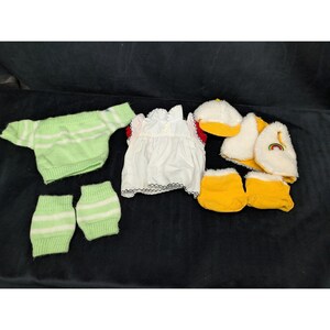 May include: A collection of doll clothing including a light green striped sweater, matching leg warmers, a white blouse with red trim, a yellow cap, a white faux fur vest with a small rainbow patch, and a pair of yellow fabric shoes.