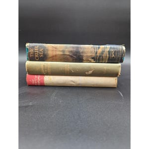 May include: A stack of three vintage hardcover books. The top book is titled The Cruel Sea by Nicholas Monsarrat. The middle book is The Heart of the Matter by Graham Greene. The bottom book has a red spine and a light tan cover.