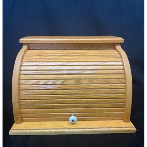 May include: A light brown wooden bread box featuring a roll-top door with horizontal slats. The unit has a flat rectangular top surface, a solid base, and a small white ceramic knob centered on the bottom slat of the sliding door mechanism.