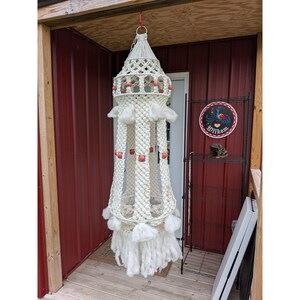 May include: A cream-colored macrame hanging lantern with a conical top and decorative wooden beads. The lantern has a fringed bottom and is suspended from a red ring. The background includes a red wall and a black metal shelf.