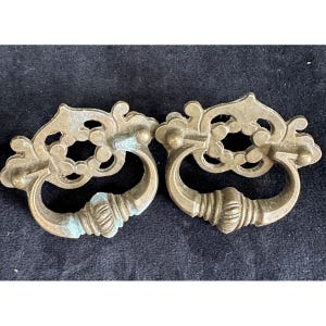 May include: Two antique brass drawer pulls with ornate, openwork backplates and curved handles. The pulls have a weathered appearance with hints of blue-green patina, suggesting age and use. The handles are approximately 4 inches wide.