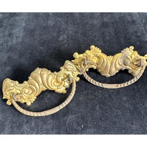 May include: Two antique brass drawer pulls with ornate, scrollwork designs. Each pull features a curved handle and intricate detailing, showcasing a vintage aesthetic. The pulls are a warm gold color, suggesting age and use.