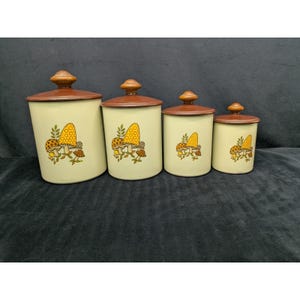May include: A set of four vintage kitchen canisters. The canisters are off-white with brown lids and wooden knobs. Each canister features a colorful mushroom design in yellow, orange, and brown. The canisters are of varying sizes.