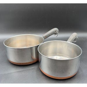 May include: Two stainless steel saucepans with copper-colored bottoms and dark gray handles. The pans are round with a slightly flared rim. The handles have a loop for hanging. The pans are empty and appear to be vintage.