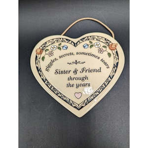 May include: Heart-shaped ceramic plaque with a cream-colored background and a decorative border. The plaque features the text "giggles, secrets, sometimes tears" and "Sister & Friend through the years". A small pink heart is also visible.