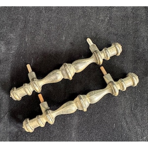 May include: Two antique brass door handles with a detailed, ornate design. The handles have a curved shape with decorative elements and a tarnished finish, suggesting age. Each handle has a central post for mounting.