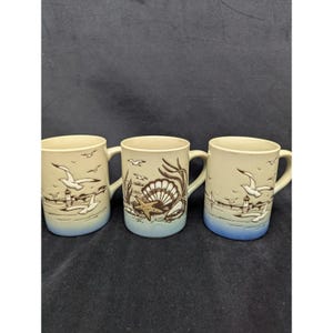May include: Three ceramic mugs with a coastal theme. Each mug features a cream-colored body with a blue base and a handle. The mugs are decorated with images of seagulls, a lighthouse, seashells, and seaweed.