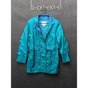 May include: Teal and blue vintage jacket with a collared neckline and button closure. The jacket features a blue lining and accents, with two front pockets and long sleeves. The jacket is hanging on a black hanger against a dark background.