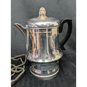 May include: A vintage electric coffee percolator made of polished chrome with a black handle and a clear glass knob on the lid. The body features a subtle etched garland design around the upper section and includes a fabric-covered power cord.