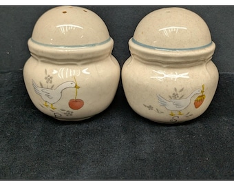 May include: A pair of speckled cream-colored ceramic salt and pepper shakers with rounded lids and blue rim accents. Each shaker features a hand-painted white goose illustration, one holding a red cherry and the other a yellow-orange fruit.