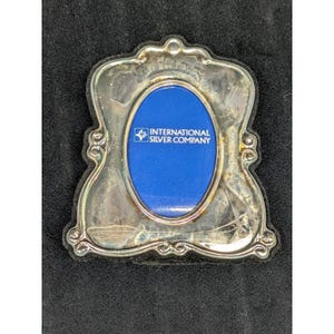May include: A silver-toned picture frame with an oval opening and ornate edges. The frame has a blue backing with the text "INTERNATIONAL SILVER COMPANY". The frame is set on a black surface.