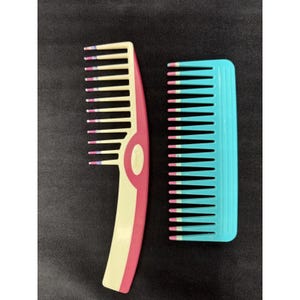 May include: Two plastic hair combs. One is a light yellow and pink comb with a curved handle. The other is a turquoise comb. Both combs have white and pink tips on the teeth. The combs are on a dark background.