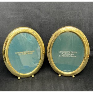 May include: Two gold-colored, oval photo frames. One is solid brass, the other is silver plated brass. Each frame has a teal-colored backing and is designed for 5x7 inch photos. The frames have a decorative leaf pattern around the edges.