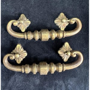 May include: Two antique brass drawer pulls with a floral design on the ends and a curved handle. The pulls are a warm gold color and appear to be made of metal. The background is a dark, textured surface.