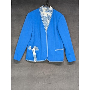 May include: A bright blue jacket with white trim and a patterned collar and pocket detail. The jacket has long sleeves and a V-neck design. The pockets have a decorative bow.