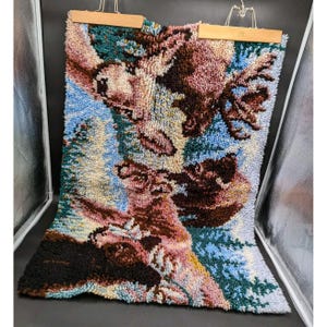 May include: A vintage, handmade rug with a detailed, textured design. The rug features a landscape scene with a variety of colors, including blue, brown, green, and pink. It is suspended from a wooden bar with metal clips.