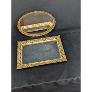 May include: Two antique gold-toned vanity trays. One is oval with a mirrored surface and ornate filigree edging. The other is rectangular with a dark surface and similar decorative filigree.