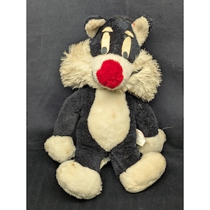 May include: Vintage plush toy cat with black fur, a white belly, and large white paws. The cat has a red nose, tan fur around the face, and large yellow eyes. A classic children's toy.