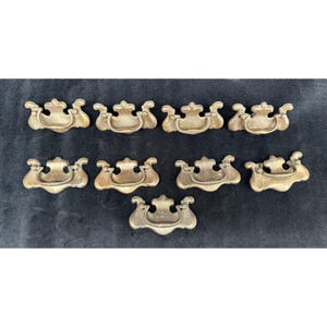 May include: A collection of ten antique brass drawer pulls. Each pull features a decorative design with a curved handle and ornate detailing. The pulls are arranged on a dark background, showcasing their aged, golden color.