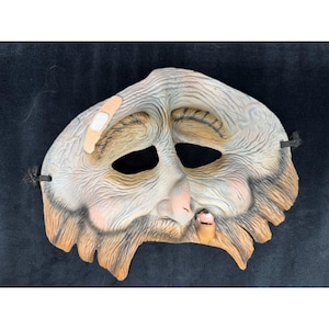 May include: A weathered mask with a sad expression, featuring a bandage on the forehead and a stubble beard. The mask has black eye holes and a string for wearing. The color palette includes shades of beige, brown, and pink.