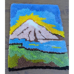 May include: A rectangular, handmade rug featuring a mountain landscape. The design includes a light pink mountain peak, a yellow sun, blue sky and water, and green and brown terrain. The rug has a textured, looped pile.