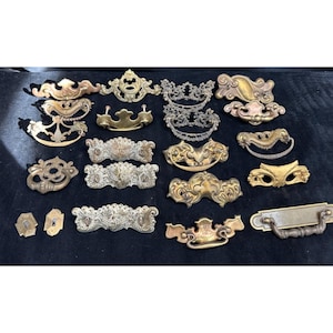 May include: A collection of antique brass drawer pulls and handles in various ornate designs. The hardware features a range of shapes, including curved, rectangular, and decorative styles, with a tarnished finish. The collection is displayed on a dark surface.