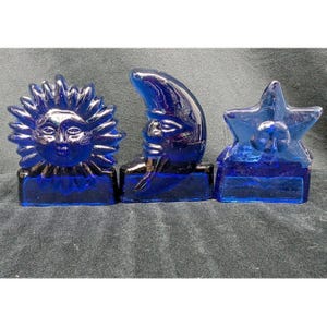 May include: Three cobalt blue glass decorative objects featuring celestial motifs. The set includes a sun with rays, a crescent moon with a face, and a five-pointed star with a face. Each piece has a flat rectangular base for stability.