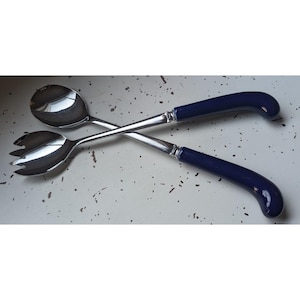 May include: A pair of salad servers featuring polished stainless steel heads and curved, solid dark blue handles. One server is a solid spoon, while the other is a fork with three tines. The two utensils are crossed over each other.
