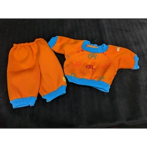 May include: A two-piece outfit consisting of orange pants and a long-sleeve shirt with blue ribbed cuffs and neckline. The shirt features a repeating pattern of the letters CPK in green, red, and orange, with a small tag on the side seam.
