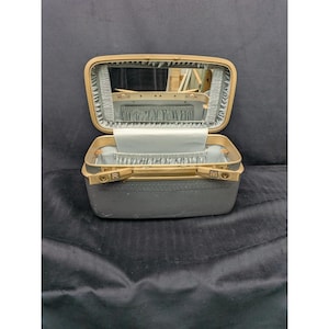 May include: An open, vintage makeup case with a gold-colored frame and a gray exterior. The interior is lined with light blue fabric and features a mirror. The case has a handle and two latches marked "R" and "M".