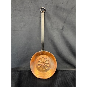 May include: A copper pan with a long, flat, dark metal handle featuring a circular hanging loop at the end. The interior of the round pan displays a stamped, recessed flower design with multiple petals centered in the base of the copper vessel.