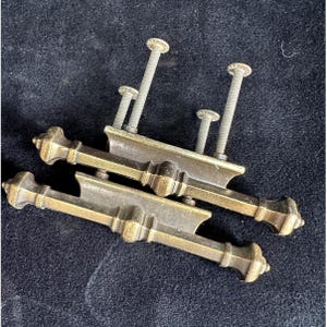 May include: Two antique brass drawer pulls with decorative detailing. Each pull has a rectangular base with ornate ends and a central design. The pulls are accompanied by several screws for installation. The hardware has a vintage aesthetic.