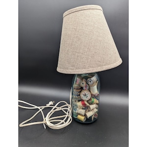 May include: A unique table lamp with a beige fabric shade. The base is a clear glass jar filled with vintage spools of thread in various colors. A white electrical cord extends from the lamp.