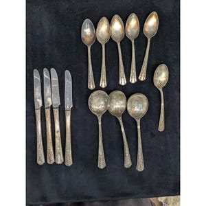 May include: A collection of twelve silver-toned metal utensils consisting of four dinner knives with straight blades and eight spoons of varying sizes, including five smaller teaspoons and three larger soup spoons, all featuring ornate decorative handles.