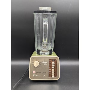 May include: Vintage Waring Blendor Custom Series 70 blender. The blender has a clear glass pitcher with a black rim, a light green base, and a control panel with a dial and buttons. The Waring logo is visible.