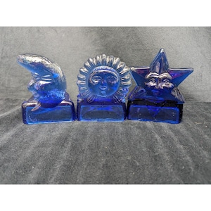 May include: Three cobalt blue glass figurines featuring celestial motifs. The set includes a crescent moon, a sun with a face, and a five-pointed star with a face. Each piece is mounted on a rectangular glass base, showcasing a translucent finish.