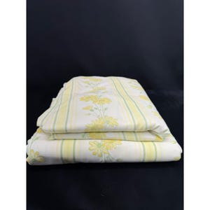 May include: Folded bedding set featuring a white background with yellow and green stripes and floral patterns. The sheets are stacked, showcasing the design. The floral pattern includes yellow daisies.